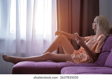 A serene blonde woman relaxes on her purple sofa near the window, wearing a silk robe. She is gently holding her smartphone, seemingly lost in thought on a peaceful morning.