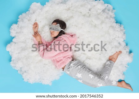 Serene calm young woman wears sleepmask on eyes comfortable pajama sleeps well sees sweet dreams poses on white fluffy cloud isolated over blue background. Top view. People and rest concept.