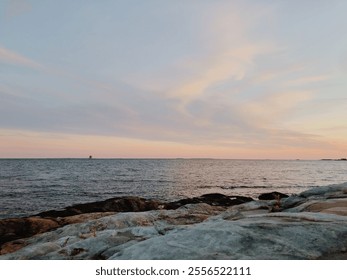 A serene coastal landscape during sunset, featuring soft pastel skies and calm ocean waves. The rocky shoreline in the foreground adds texture to the tranquil scene. - Powered by Shutterstock