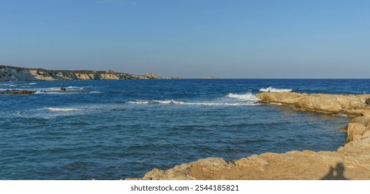 A serene coastal landscape features sparkling blue waters and soft waves against sunlit rocks at dusk. - Powered by Shutterstock