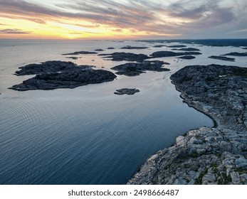 A serene coastal landscape featuring rocky shores, calm waters, and a colorful sky at sunset. The foreground includes scattered stones and greenery, while distant islands are visible on the horizon. - Powered by Shutterstock