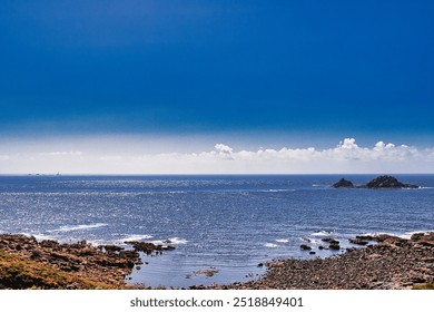 A serene coastal landscape featuring rocky shores, calm blue waters, and a clear sky with a few clouds. At Cape Cornwall, UK. - Powered by Shutterstock