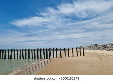 A serene coastal landscape featuring a sandy beach with weathered wooden groynes extending into the gentle waves. the bright blue sky is adorned with wispy clouds, and a rocky breakwater lines - Powered by Shutterstock