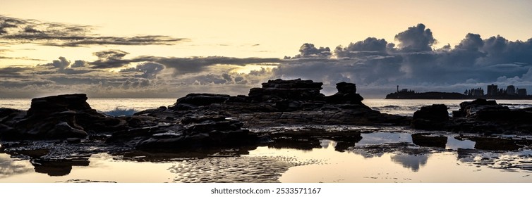 A serene coastal landscape at sunrise with rock formations and reflections in the water, with a city skyline in the distance. - Powered by Shutterstock