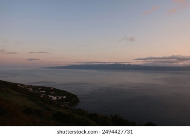 A serene coastal scene at dusk featuring a calm sea, silhouetted Pico island in the distance, and a small village nestled on a green hillside. São Jorge island, Azores, Portugal. - Powered by Shutterstock