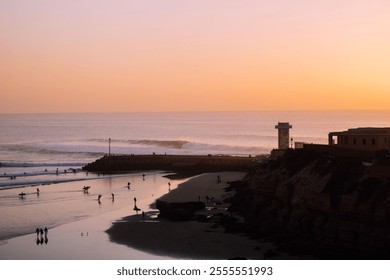 A serene coastal sunset with surfers in the water, gentle waves breaking on the shore, and a lighthouse overlooking the beach bathed in warm orange hues. - Powered by Shutterstock