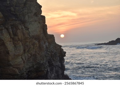 A serene coastal sunset at Ussangoda beach, Sri-Lanka, with a rugged cliff in the foreground, calm waves, and a glowing sun on the horizon. - Powered by Shutterstock