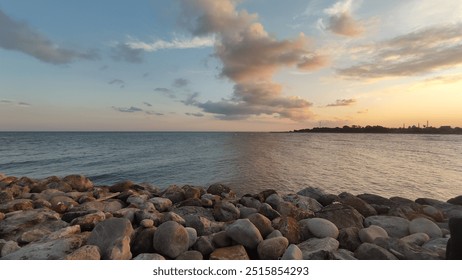 Serene coastal view with calm blue waters and scattered clouds under a pastel sunset sky, rocky shore in the foreground, offering a peaceful and tranquil scene - Powered by Shutterstock