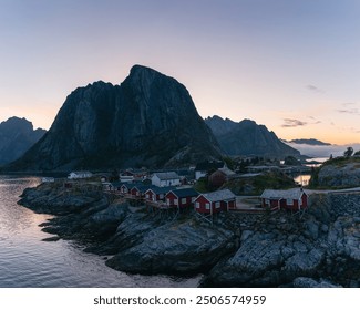 A serene coastal village in Lofoten, with traditional red and white houses, sits on rocky shores, backed by towering, rugged mountains at dusk. The calm waters reflect the tranquil, pastel-colored sky - Powered by Shutterstock