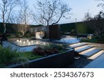 Serene courtyard with a sleek metal sculpture, rippling water feature, manicured greenery, and ambient lighting under a twilight sky.