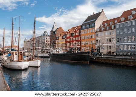 A serene day at Nyhavn, Copenhagen, with boats moored in the canal, reflecting the colorful facades of European townhouses, under a blue sky with scattered clouds.