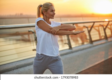 Serene dreamy athletic suntanned blonde woman performing a side arm stretch on a pedestrian bridge - Powered by Shutterstock