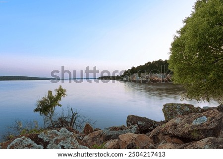 A serene dusk scene at Atoka Oklahoma Park, showcasing the calm lake waters reflecting the soft hues of the sky. The rocky shoreline and dense tree line create a peaceful and scenic environment 