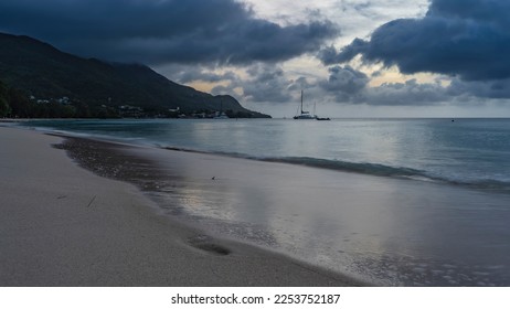 A serene evening on a tropical island. Ocean waves roll onto the beach. Wet sand glistens. Yachts on the horizon. A green hill against the sky and clouds. Shades of blue. Seychelles. Mahe. Beau Vallon - Shutterstock ID 2253752187