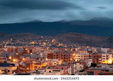 A serene evening view of a densely populated city nestled against rolling hills, under the subtle, soft glow of streetlights illuminating the architectural landscape. The buildings, painted in warm hu - Powered by Shutterstock