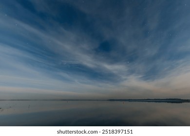 A serene, expansive lake reflects the soft glow of a starry night sky. Wispy clouds stretch across the horizon, adding to the calm, tranquil atmosphere. The distant shoreline is faintly visible - Powered by Shutterstock