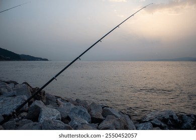 Serene Fishing by the Seaside at Dusk: A Quiet Moment with a Fishing Rod Overlooking the Calm Waters and Rocky Shore - Powered by Shutterstock