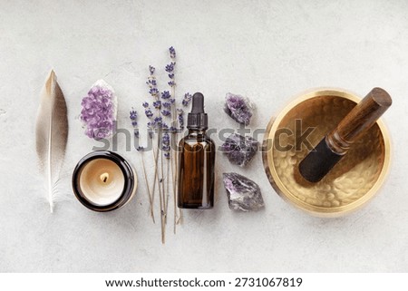 A serene flat lay composition featuring a variety of spiritual and wellness tools, including a brown glass essential oil dropper bottle, dried lavender sprigs, raw and polished amethyst crystals, a feather, a lit candle, a brass singing bowl with wooden mallet, sage smudge stick, and palo santo.