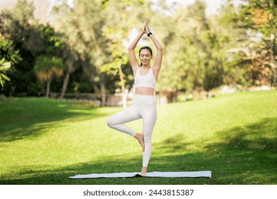 Serene glad slim caucasian woman practicing the Tree Pose in yoga, balancing on one leg with hands in prayer position above head on a mat in a tranquil park setting, outside - Powered by Shutterstock