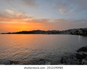 A serene Greek sunset over the Aegean Sea, with orange and pink hues reflecting on calm waters. Silhouetted hills and a coastal village complete the tranquil scene - Powered by Shutterstock