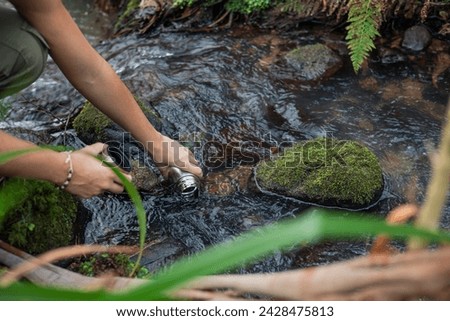 In the serene heart of the forest, skilled hands dip into a crystal-clear river, gathering water in a metal container. This image captures the essence of human connection with nature, 