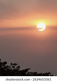 A serene image captures the sun setting through a hazy sky, casting warm orange and pink hues. Silhouetted trees are visible at the bottom. A peaceful end-of-day scene. #Sunset #HazySunset #WarmLight 