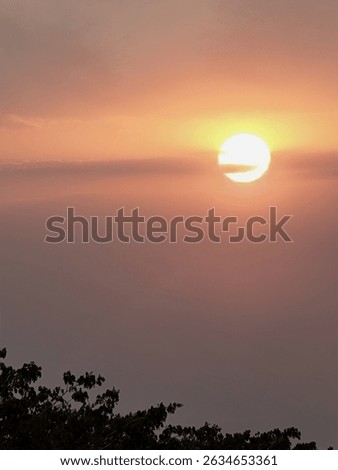 A serene image captures the sun setting through a hazy sky, casting warm orange and pink hues. Silhouetted trees are visible at the bottom. A peaceful end-of-day scene. #Sunset #HazySunset #WarmLight 