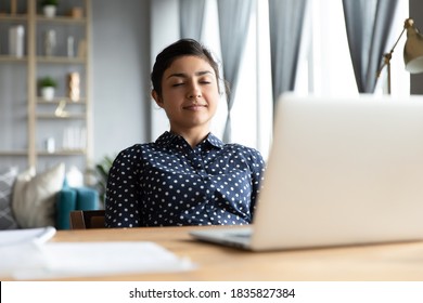 Serene indian business woman smiling sit at desk lean on office chair, enjoy break pause after working hard closing eyes daydreaming relaxing, feels satisfaction after project accomplishment concept