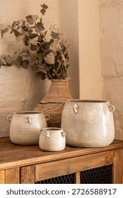 A serene interior decor scene featuring three handcrafted cream ceramic pots with side handles, arranged on a wooden table next to a large woven straw vase with dried eucalyptus branches against a whi