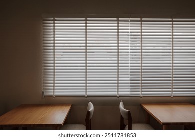 A serene café interior featuring two wooden tables and matching upholstered chairs positioned in front of large windows with closed blinds.  - Powered by Shutterstock