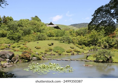 A serene Japanese garden features a pond with lily pads and a stepping stone path crossing the water. Lush greenery surrounds the scene, and a traditional wooden structure peeks through the trees unde - Powered by Shutterstock