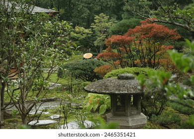 A serene Japanese garden with a stone lantern, lush greenery, and a person wearing a traditional hat. The garden features various plants, including trees with red leaves, and a stone pathway. - Powered by Shutterstock