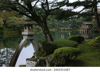 A serene Japanese garden with a stone lantern, lush greenery, and a calm pond reflecting the surrounding trees. A traditional wooden building is visible in the background. - Powered by Shutterstock