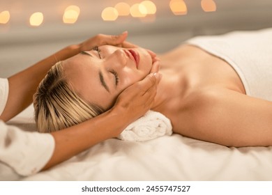 A serene lady lies on her back, eyes closed, with a peaceful expression as she receives a facial massage. Soft lighting and blurred lights in the background create an atmosphere of tranquility