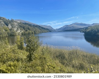 A serene lake with calm water reflecting a blue sky, surrounded by lush greenery, rocky hills, and distant mountains under bright sunlight, creating a peaceful, natural landscape. - Powered by Shutterstock