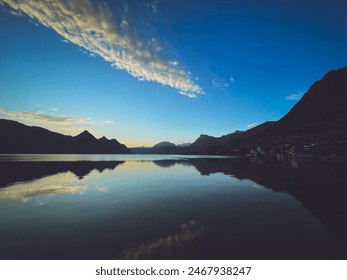 A serene lake with calm waters reflecting the surrounding mountains at either sunrise or sunset. A dock with wooden posts extends into the water. The sky is clear with scattered clouds - Powered by Shutterstock