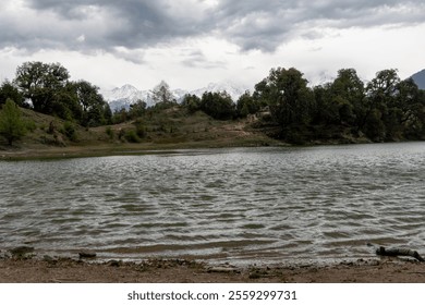 A serene lake with gentle ripples reflects the cloudy sky, surrounded by dense green trees and distant snow-capped mountains. Peaceful overcast atmosphere at Deoria tal lake trek in Ukhimath, India. - Powered by Shutterstock