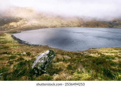 A serene lake lies nestled among rolling hills, enveloped in soft morning mist, creating a peaceful atmosphere in a remote countryside location. - Powered by Shutterstock