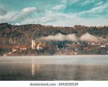 A serene lake with a mist-covered surface reflects the colorful autumn trees on the hillside, where a small village nestles among the foliage - Powered by Shutterstock
