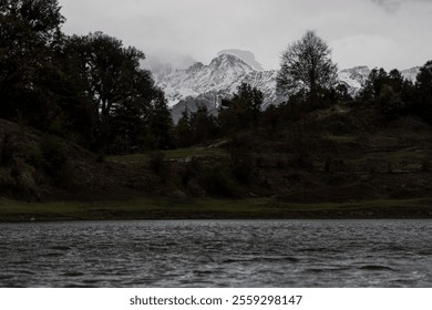 A serene lake with rippling water at the foreground, surrounded by rugged terrain and dense trees. Snow-capped mountains rise dramatically in the background under a dark and cloudy sky at deoria lake. - Powered by Shutterstock