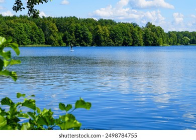 A serene lake scene featuring a lone paddleboarder gliding across the calm waters, surrounded by lush green forests under a clear blue sky. Captured in summer, this image portrays tranquility  - Powered by Shutterstock