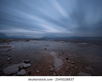 Serene lake shore at dusk with cloudy sky and distant mountains. Calm waters and muted colors create a tranquil, moody atmosphere, ideal for themes of nature, peace, and solitude - Powered by Shutterstock