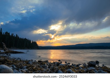 A serene lake at sunset with a rocky shore in the foreground. The sky is partially clouded with the sun peeking through, casting warm hues across the landscape. Forested hills and trees border the lak - Powered by Shutterstock