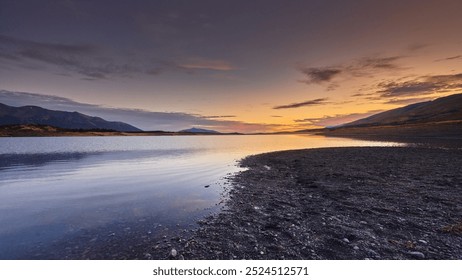 a serene lakeside at dusk with a glowing orange sky, dark mountains in the distance, and calm waters reflecting the fading light. Lago  Roca, Patagonia, Argentina - Powered by Shutterstock