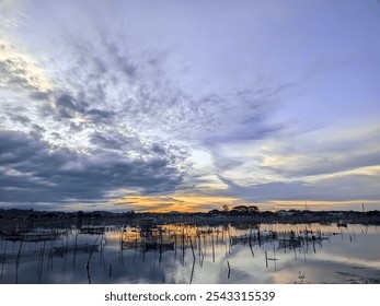 A serene lakeside landscape during sunset, with dramatic clouds reflected in calm waters. Sticks are arranged throughout the lake, possibly used for fishing nets or other traditional water activities  - Powered by Shutterstock