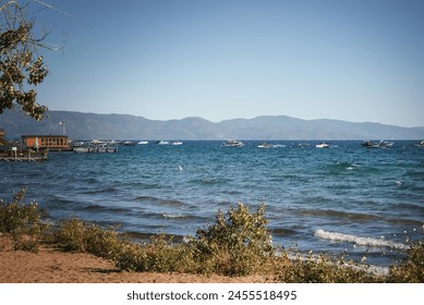 Serene lakeside scene with clear blue sky, sandy beach, green bushes, lake water with boats, wooden dock, American flag, mountains in the background. Ideal for outdoor activities in the United States. - Powered by Shutterstock