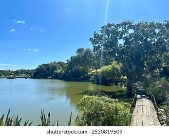 A serene lakeside scene with a clear blue sky, lush green trees, and a charming red gazebo in the distance. The wooden bridge crossing over part of the lake leads to the far side of the scene. - Powered by Shutterstock