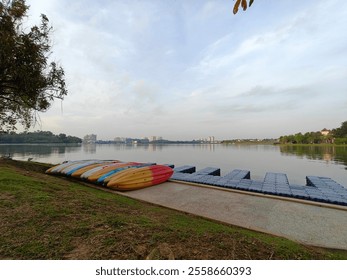 A serene lakeside scene with colorful kayaks lined up on the shore, a floating dock extending into the calm water, and distant buildings under a partly cloudy sky. - Powered by Shutterstock