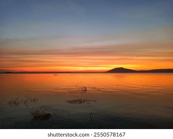 A serene lakeside sunset. A small boat is silhouetted against the horizon, and the silhouette of a mountain range can be seen in the distance. The overall  atmosphere is peaceful and tranquil. - Powered by Shutterstock