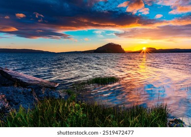 A serene lakeside sunset with vibrant colors in the sky, reflected on the calm water. The horizon features a silhouetted island and rolling hills. In the foreground, there is a shoreline with grass an - Powered by Shutterstock